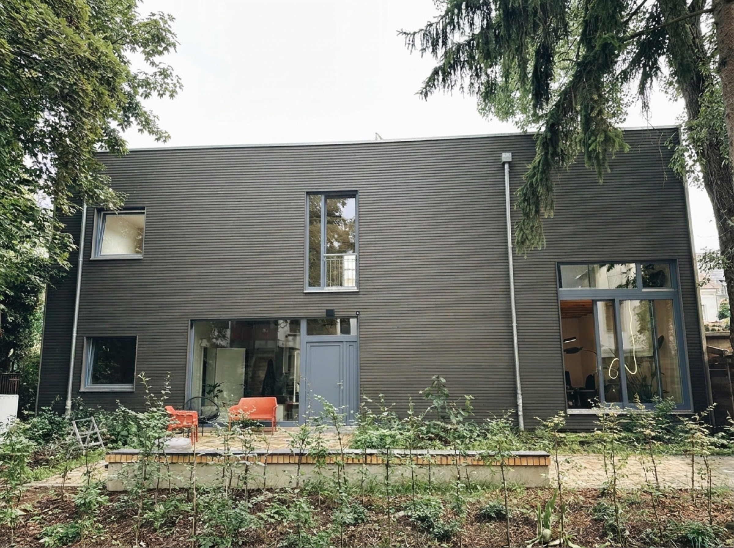 Das tribehouse von außen: dunkle Holzfassade, große Fenster, Hinterhof mit Bäumen